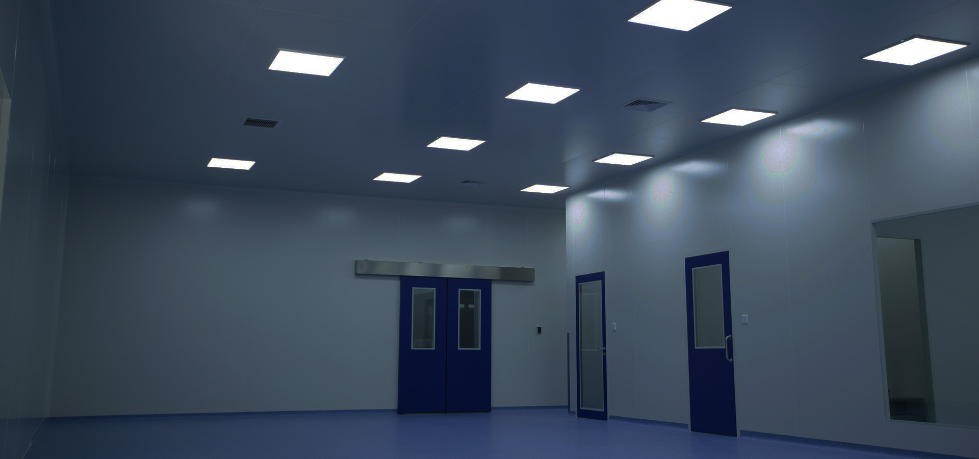 Interior de sala limpia con paneles murales claros, puertas azules y luces de techo